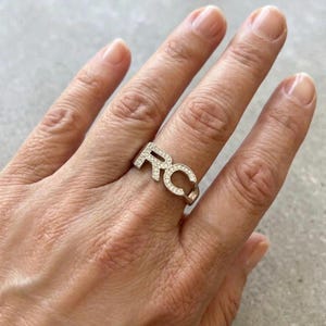 Puede incluir: Un anillo plateado con las letras "RC" incrustadas con pequeñas piedras preciosas transparentes. El anillo está en un dedo, mostrando el diseño y el brillo de la joya.