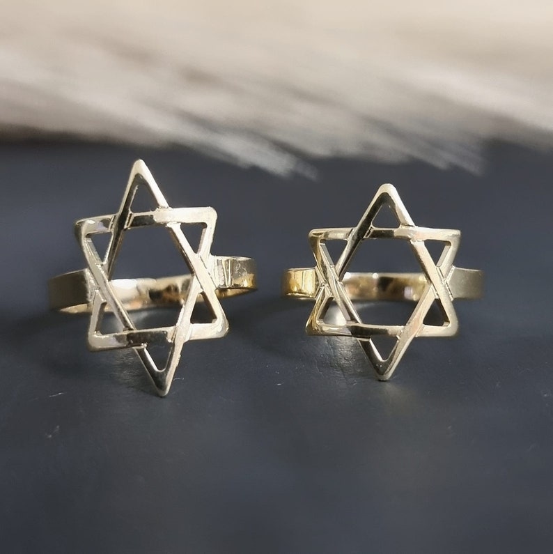 May include: Two gold-colored rings, each featuring a Star of David design. The rings are set against a dark background, with the six-pointed stars prominently displayed. The rings appear to be made of a shiny metal.