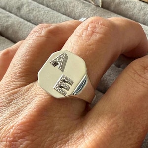 Peut inclure: Une bague chevalière en argent avec les lettres "A" et "E" incrustées de petites pierres scintillantes. La bague a une face octogonale polie et un anneau lisse et arrondi. La bague est montrée sur un doigt.