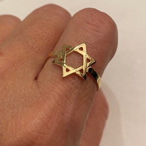 May include: A gold-colored ring featuring a Star of David design. The ring is made of a shiny, reflective metal and is worn on a finger. The ring's design is a six-pointed star, a symbol of Judaism.