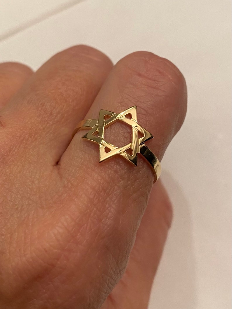 May include: A gold-colored ring featuring a Star of David design. The ring is made of a shiny, reflective metal and is worn on a finger. The ring's design is a six-pointed star, a symbol of Judaism.