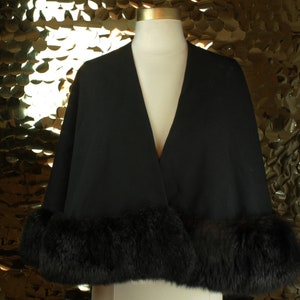 May include: A black cape with a fur trim around the edges. The cape is displayed on a mannequin against a gold sequin background.