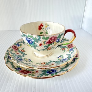 May include: A vintage teacup and saucer set. The ivory-colored cup and saucer feature a floral pattern in red, blue, and pink, with gold trim. The cup has a red handle. The set is designed for tea service.