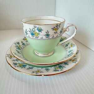 May include: A vintage teacup, saucer, and side plate set. The cup and saucer are light green with floral patterns in blue and yellow. The rims are trimmed with gold.