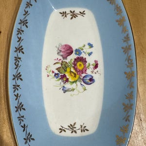 May include: An oval-shaped, light blue serving platter with a white center. The center features a floral bouquet with pink, purple, yellow, and blue flowers. Gold filigree and floral accents adorn the edges.