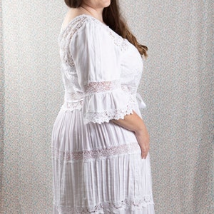 Op de afbeelding: Een witte maxi-jurk met kantdetails en een gelaagde rok. De jurk heeft een ronde hals en 3/4 mouwen.
