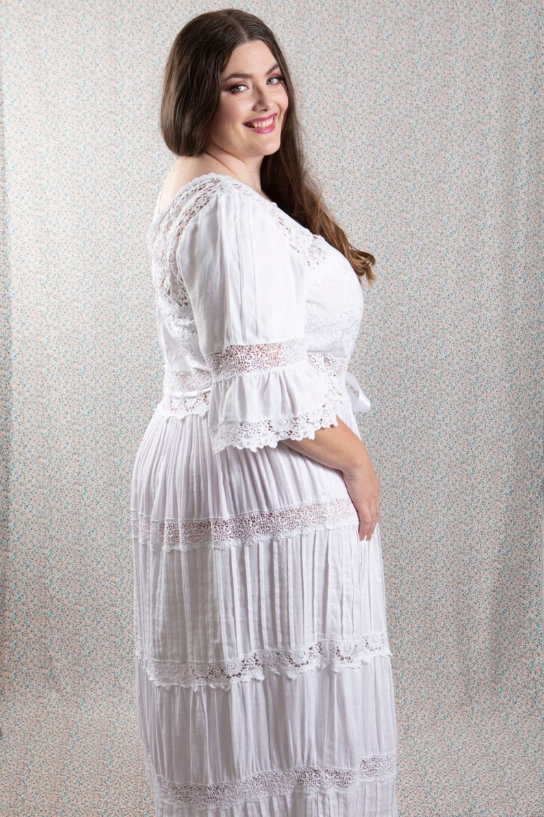 Op de afbeelding: Een witte maxi-jurk met kantdetails en een gelaagde rok. De jurk heeft een ronde hals en 3/4 mouwen.