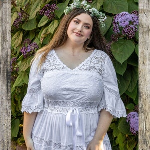 Op de afbeelding: Een vrouw die een witte kanten jurk draagt met een wit lint dat in de taille is geknoopt. Ze staat voor een groene, bladerige achtergrond met paarse bloemen.