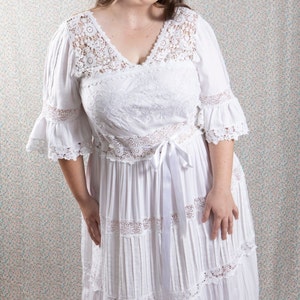 Op de afbeelding: Een witte maxi-jurk met een kanten overlay en een satijnen lint strik aan de taille. De jurk heeft een v-hals en een gelaagde rok.