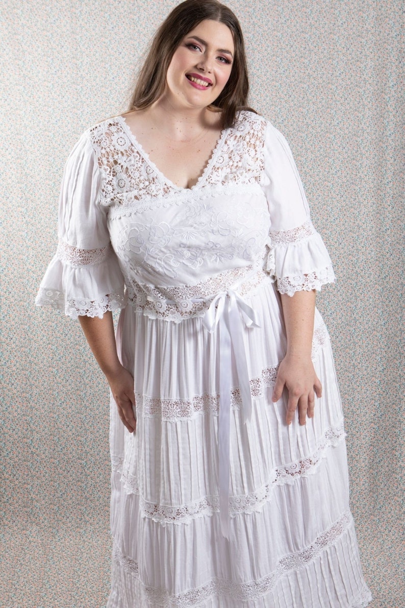 Op de afbeelding: Een witte maxi-jurk met een kanten overlay en een satijnen lint strik aan de taille. De jurk heeft een v-hals en een gelaagde rok.