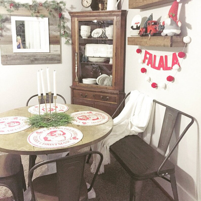 May include: A round wooden table with four black metal chairs and a white throw blanket on a black metal bench. The table is set with red and white Santa Claus themed placemats and a green garland with a gold candle holder in the centre. A wooden hutch with glass doors is in the background, filled with white dishes. A red and white pom-pom garland with the word "FALALA" in red letters hangs on the wall.