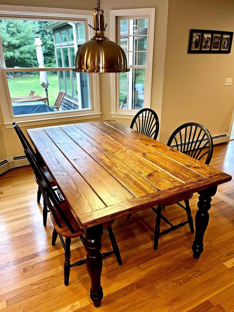 May include: A rustic wooden dining table with a natural wood finish and black turned legs. The table is surrounded by four black chairs with wooden seats. The table is in a room with hardwood floors and a window in the background.