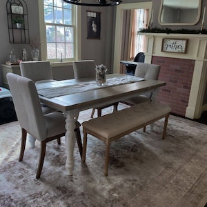 May include: A farmhouse style dining room with a white and gray table, four chairs, and a bench. The table has a whitewashed finish and turned legs. The chairs are upholstered in a gray fabric. The bench is upholstered in a beige fabric. The room has a gray rug with a distressed pattern. The room also has a fireplace with a brick surround and a mirror above it. The fireplace has a sign that says "gather" on it.