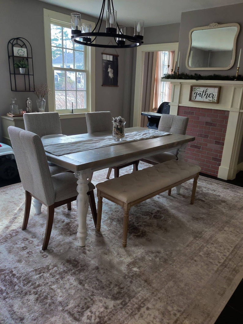 May include: A farmhouse style dining room with a white and gray table, four chairs, and a bench. The table has a whitewashed finish and turned legs. The chairs are upholstered in a gray fabric. The bench is upholstered in a beige fabric. The room has a gray rug with a distressed pattern. The room also has a fireplace with a brick surround and a mirror above it. The fireplace has a sign that says "gather" on it.