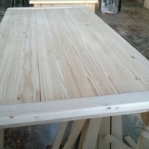 May include: A large, unfinished wooden table top with a light wood grain. The table top has a simple, rectangular shape and is made of several planks of wood.