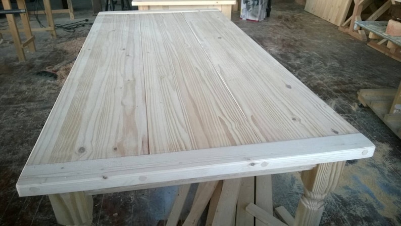 May include: A large, unfinished wooden table top with a light wood grain. The table top has a simple, rectangular shape and is made of several planks of wood.