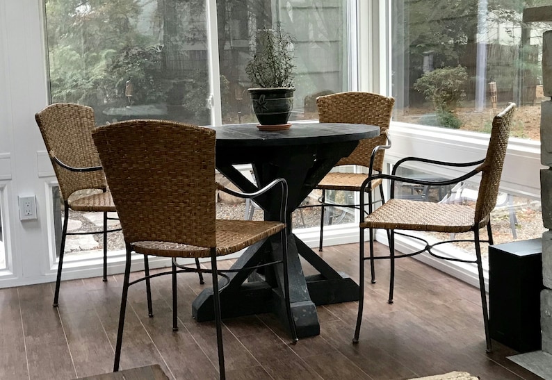 May include: A black wooden table with a cross-shaped base and four wicker chairs with black metal frames. The table is set in a sunroom with large windows and a potted plant on the table.