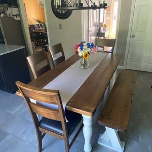 May include: A wooden dining table with a white base and a matching bench. The table is set with a white tablecloth and a vase of red, white, and blue flowers.