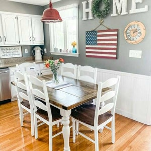 May include: A white farmhouse dining table with a wooden top and white turned legs. The table is surrounded by six white chairs with wooden seats. The table is set with a vase of flowers and a placemat. The wall behind the table is painted gray and features a wooden sign that says "HOME" with a green wreath hanging above it. An American flag hangs on the wall to the right of the sign.