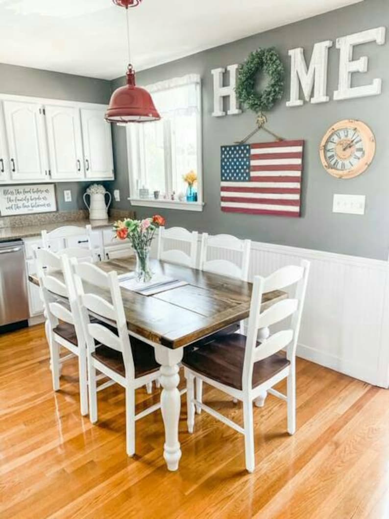 May include: A white farmhouse dining table with a wooden top and white turned legs. The table is surrounded by six white chairs with wooden seats. The table is set with a vase of flowers and a placemat. The wall behind the table is painted gray and features a wooden sign that says "HOME" with a green wreath hanging above it. An American flag hangs on the wall to the right of the sign.