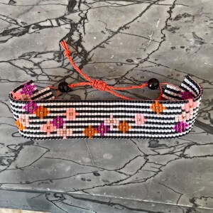 May include: A beaded bracelet with a black and white striped pattern, accented with pink, orange, and purple floral designs. The bracelet is connected by an orange cord with black beads.