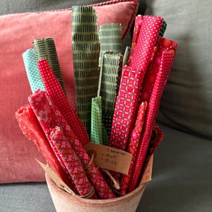 Peut inclure: Un assortiment d'emballages alimentaires en cire d'abeille colorés dans un petit pot en terre cuite. Les emballages sont roulés et présentent divers motifs rouges, verts et bleus. Une petite étiquette est attachée avec du texte manuscrit.