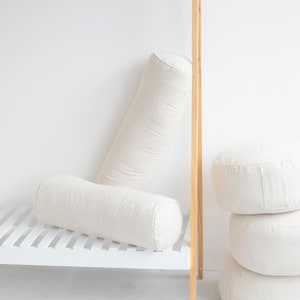May include: Two white cylindrical pillows, one lying on a white shelf and the other standing upright against a white wall. Three more white round pillows are stacked on top of each other on the floor.