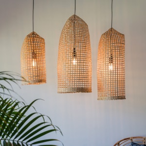 May include: Three woven wicker pendant lights hang from the ceiling in a room with a white wall. The lights are lit and cast a warm glow. A wicker chair with a white cushion and a blue patterned pillow sits in the room. A wicker side table with a book on top sits in the foreground.