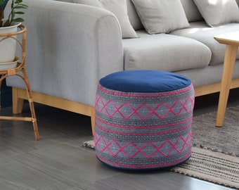 Puede incluir: Un puf redondo de color azul y gris con estampado y bordado rosa. El puf está sobre una alfombra gris delante de un sofá gris.