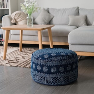 Può includere: Un pouf rotondo blu e bianco con un design geometrico. Il pouf è seduto su un tappeto di fronte a un divano grigio.