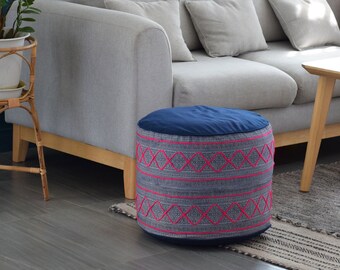 Puede incluir: Un puf redondo azul y gris con detalles bordados en rosa. El puf está sobre una alfombra gris y blanca delante de un sofá gris.
