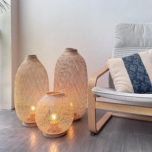 May include: Three woven wicker lamps with warm light shining through the intricate pattern. The lamps are on a gray tiled floor in front of a light wood chair with a white cushion.