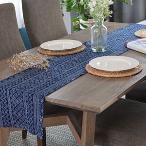 May include: A wooden dining table with a blue and white patterned table runner, two white plates with woven placemats, and a vase of white flowers.