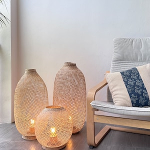 May include: Three woven bamboo lamps with warm white light bulbs. The lamps are different sizes and are sitting on a light brown tiled floor. A light gray chair with a white pillow is in the background.