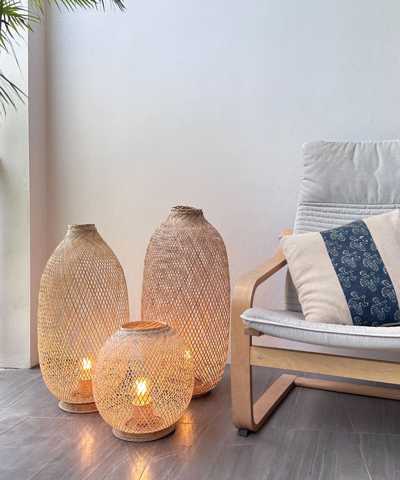 May include: Three woven bamboo lamps with warm white light bulbs. The lamps are different sizes and are sitting on a light brown tiled floor. A light gray chair with a white pillow is in the background.