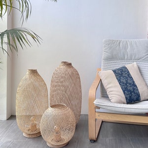 May include: Three woven wicker lampshades of varying sizes, with the largest one standing on the floor and the two smaller ones sitting on the floor in front of it. The lampshades are made of natural woven material and have a light brown color. A light brown wooden chair with a light gray cushion and a blue and white patterned pillow is in the background.