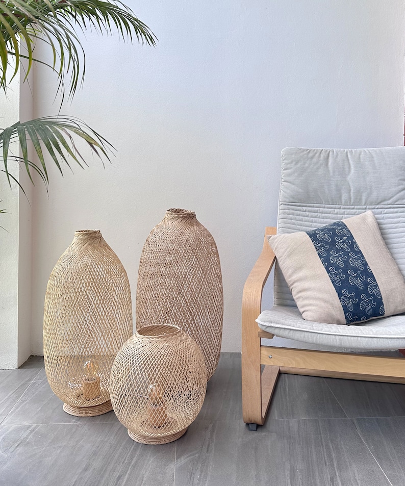 May include: Three woven wicker lampshades of varying sizes, with the largest one standing on the floor and the two smaller ones sitting on the floor in front of it. The lampshades are made of natural woven material and have a light brown color. A light brown wooden chair with a light gray cushion and a blue and white patterned pillow is in the background.
