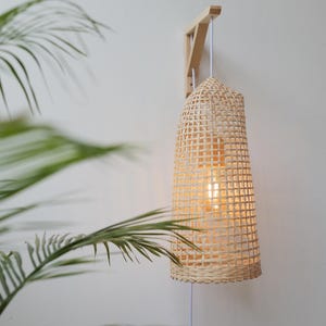 Op de afbeelding: Een geweven bamboe wandlamp met een gloeilamp erin, hangend aan een houten beugel. De lamp heeft een cilindrische vorm met een rasterpatroon. Een wit snoer hangt naar beneden. De achtergrond is een witte muur met groene palmbladeren.