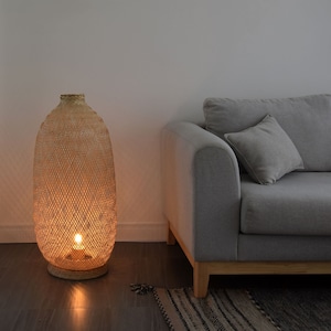 May include: A woven wicker lampshade with a warm light illuminating the interior. The lamp is placed on a wooden floor next to a gray sofa.