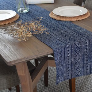 May include: A blue and white patterned table runner with a geometric design. The runner is laid on a wooden table with two place settings and a bouquet of dried flowers.