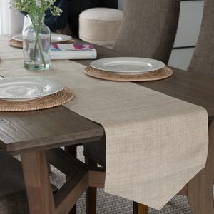 May include: A beige linen table runner with a subtle textured pattern. The runner is laid on a dark wood table with two white plates and woven placemats.