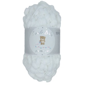 May include: A skein of white pompom yarn labeled "Zarela Baby." The yarn is fluffy and textured, with small, round pompoms. The label features a teddy bear illustration and the words "pompom yarn."