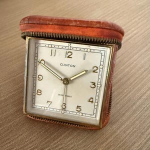 May include: A vintage Clinton travel alarm clock in a brown leather case. The square clock face has gold numerals and hands, with the word "Clinton" above the center. The case has a zipper closure.