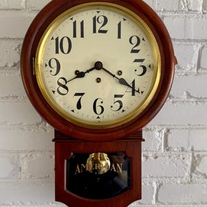 May include: A round wooden wall clock with a brass bezel and a cream-colored face featuring black Arabic numerals. Below the clock face is a rectangular wooden section with a glass panel displaying the word AMERICAN and a gold pendulum.