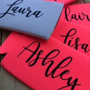 May include: A close-up of six personalized pink and light blue neoprene can coolers with names written in black. The names are Laura, Kim, Blair, Lisa, and Ashley.