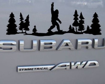 May include: Black vinyl sticker of a Bigfoot figure with raised arms and a peace sign, walking through a forest of pine trees. The sticker is on the back of a silver Subaru with the text "SUBARU" and "SYMMETRICAL AWD" on the back.