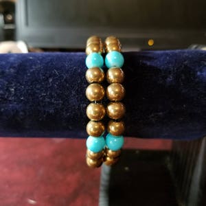 May include: Two bracelets made of gold beads with turquoise accents. The bracelets are stacked on top of each other.