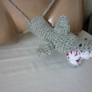 May include: A gray crocheted shark puppet with a white and red open mouth. The puppet is hanging from a string.