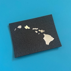 May include: A black square with a textured surface features a metallic silver outline of the Hawaiian Islands. The islands are in a polished silver finish, contrasting with the dark background. The square is centered on a bright blue background.
