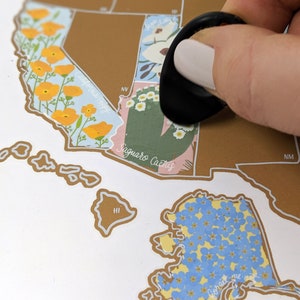 May include: A gold-colored scratch-off map of the United States with a black finger covering the state of Arizona. The state of Arizona is labeled "Saguaro Cactus".